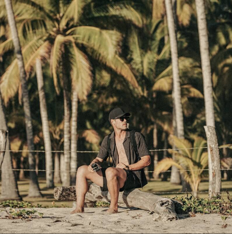 Man sitting on a log by palm trees on a beach